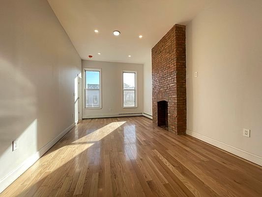 Empty room with wooden floors, a brick accent wall, and two windows. Ceiling lights are on, and sunlight streams in.