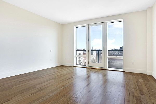 Empty room with hardwood floors, white walls, and large windows overlooking a cityscape.