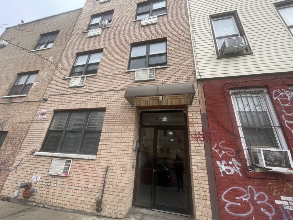 A four-story brick building with multiple windows, air conditioning units, and graffiti on the front wall.