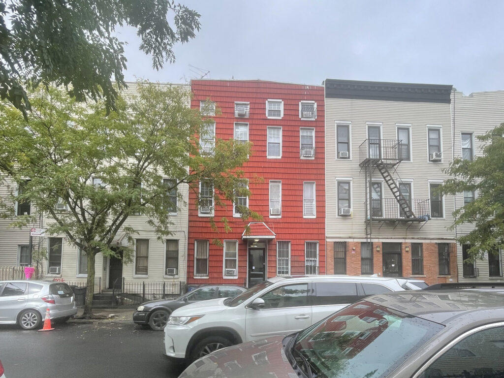 Street view of a row of three-story buildings with parked cars in front; one building is painted red, the others are gray.