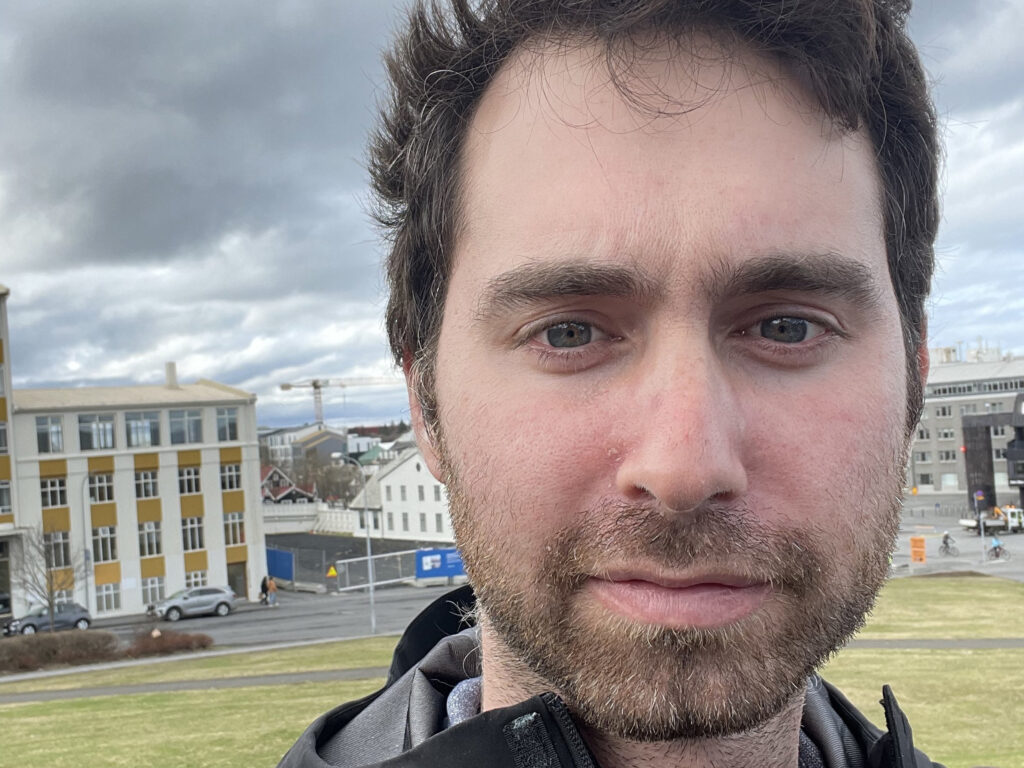 A man with short hair and a beard stands outdoors with buildings and a cloudy sky in the background.