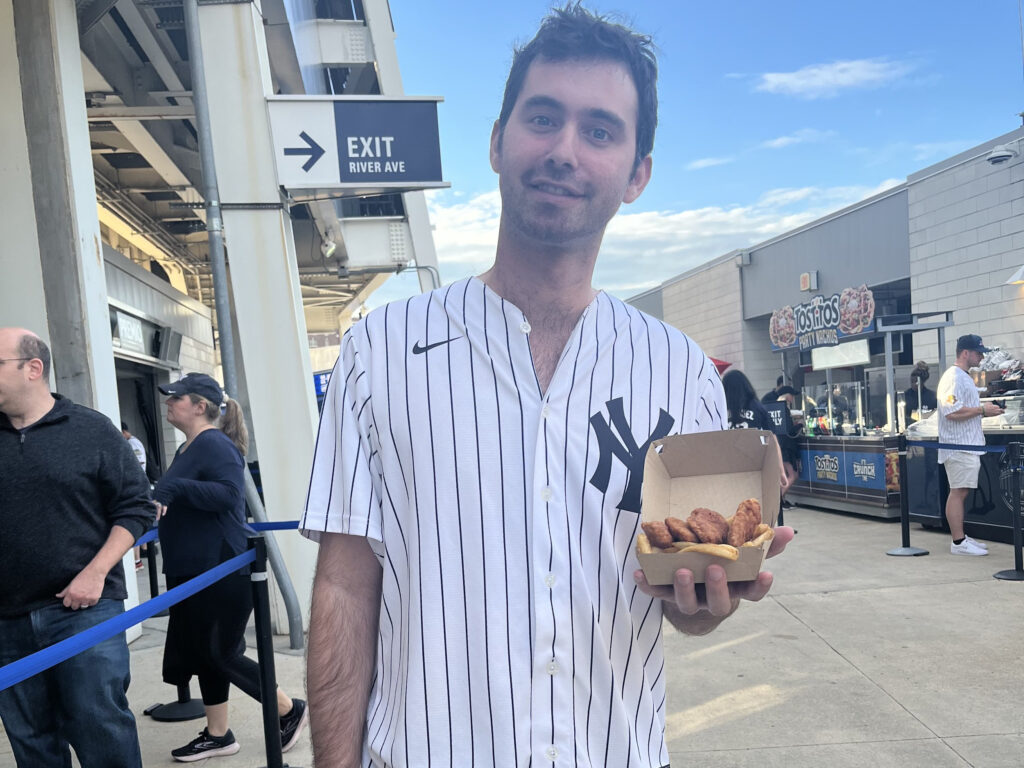 A person in a Yankees jersey holds a box of food outside a stadium near an "Exit" sign.