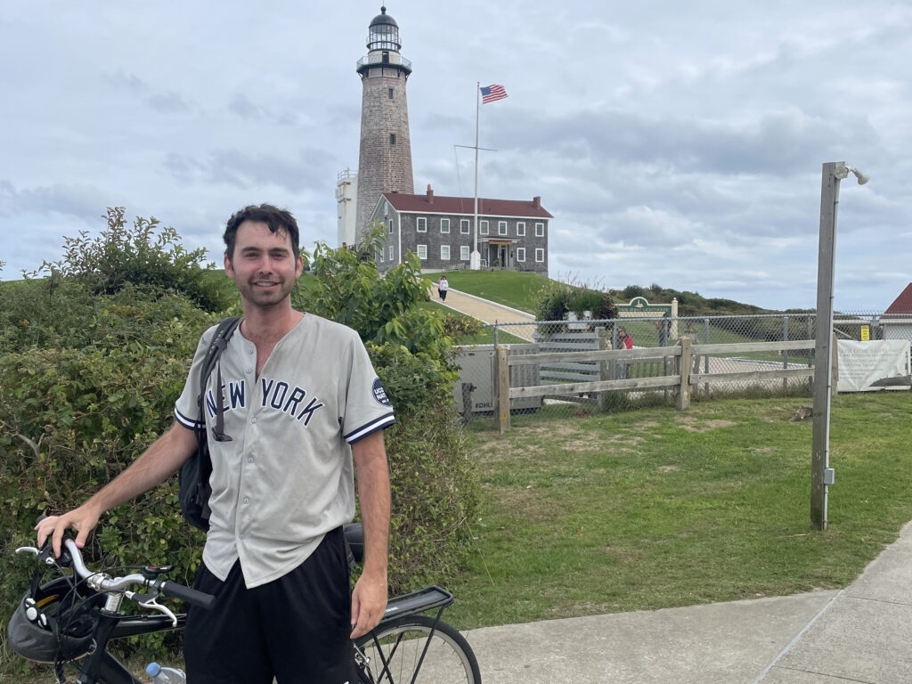 Man in a New York shirt stands with a bicycle near a lighthouse and a building with a U.S. flag on a cloudy day.